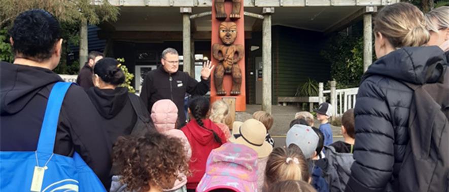 A man speaking to children and adults in front of the Arataki pou.