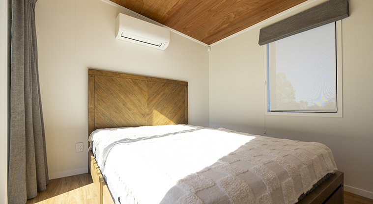 Te Kopua tiny home - Bedroom with heatpump on the wall above the bed and a window with a blind.