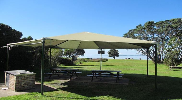 Cornwallis, Waitākere Ranges Regional Park (Pūponga Peninsula) - Section of the site showing a large umbrella over picnic tables and a barbecue.