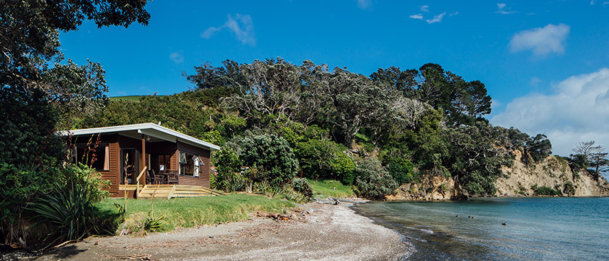 Graham Bach, Scandrett Regional Park, Auckland small 70's style house on a sweeping shoreline with cliffs and Pohutukawa trees in the background. Showcasing the type of accommodation we offer.