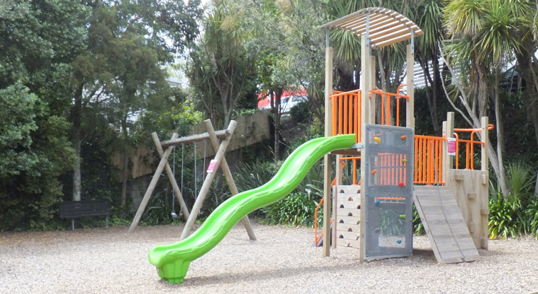 Aitken Reserve - Play module with climbing walls, platforms and a bright green slide. The set of two swings and trees are in the background. Photo credit: J Grigg.