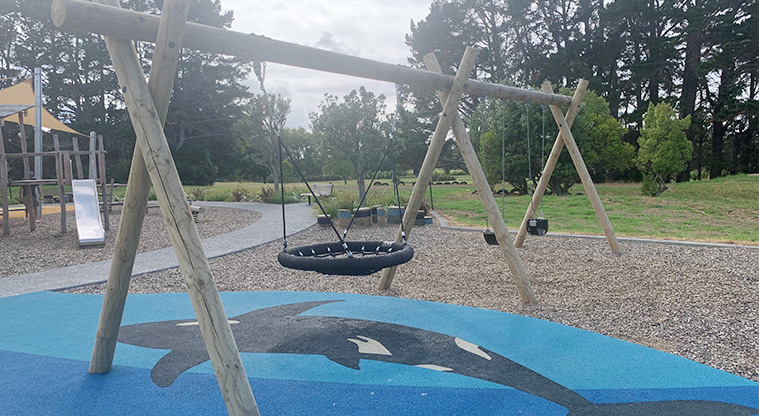 Ngahere Ki Uta / Clarks Beach Recreational Reserve and Golf Club - Swing set with a basket swing, a belt swing and a bucket swing, with the golf course and trees in the background.