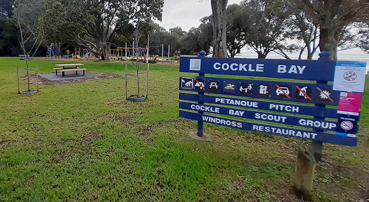 Cockle Bay Reserve - Sign at the entrance showing what is included within the reserve, with picnic tables, trees and the playground in the background.