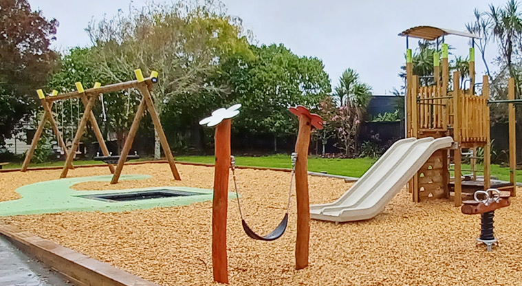 Collins Park - Junior play module with a double low slide, swing set in the background, and a low sling swing and rocker toy in the foreground.
