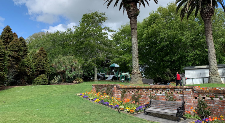 David Nathan Park - Section of the lawn and gardens with a bench in front of the wall and the playground in the background.