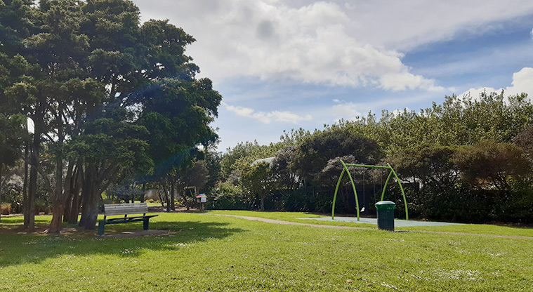 Francis Reserve - Grassed area with a seat under a large tree, and the swings in the background. Photo credit: T Hodder.