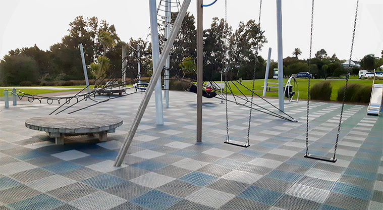 Heron Park - Playground with swings, climbing structure, bucket swing, slide, wooden seat and spinning bucket. Car park and picnic bench in the background.