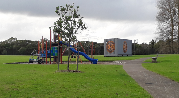 Imrie Park - Colourful playground with slide, climbing equipment and swings with open grassed space, trees and a large container in the background.