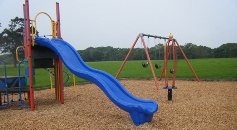 Imrie Park - Blue slide with a set of three swings, open grassed space and trees in the background.