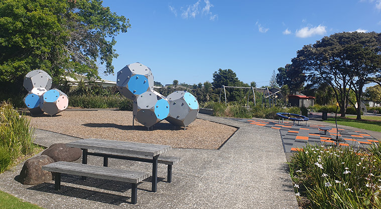 Kuaka Park - Lower playground with a picnic table, climbing shapes and spinning toys with a path down the middle and trees in the background. Photo credit: J Grigg.