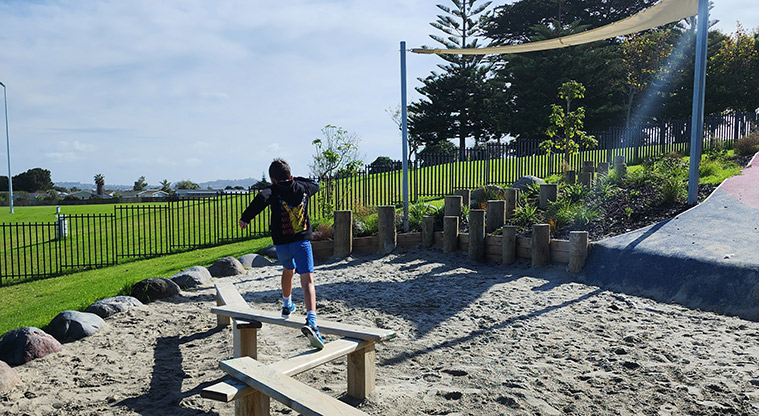 Ngā Hau Māngere / Māngere Centre Park - Child jumping across the balance beams and a section of garden with the playground fence and open grassed space in the background.