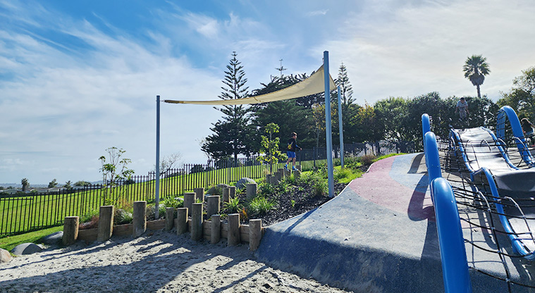 Ngā Hau Māngere / Māngere Centre Park - Blue waterfall-like climbing structure running down the hill, garden surrounded by post stumps and shade sail over the garden.