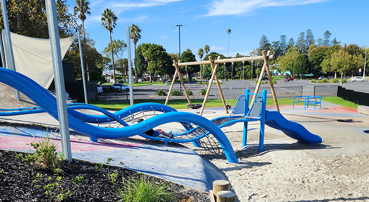 Ngā Hau Māngere / Māngere Centre Park - Section of the blue waterfall-like climbing structure with the swings, carousel, fence and car park in the background.