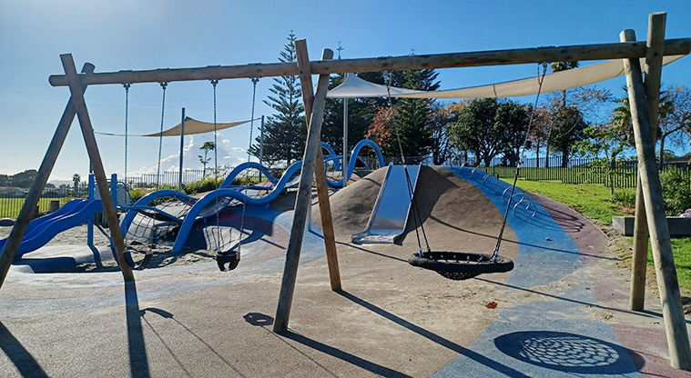 Ngā Hau Māngere / Māngere Centre Park - Set of three swings (including a basket swing) with a slide and the waterfall-like climbing structure in the background.
