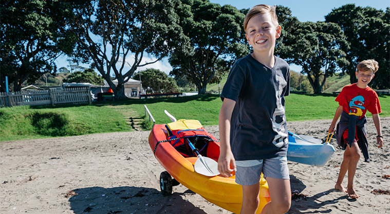 Martins Bay Holiday Park - Two children walking across sand pulling a canoe and a kayak.