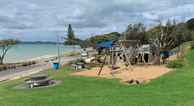 Omana Esplanade - The playground with a picnic table, the road and view of the Hauraki Gulf Islands.