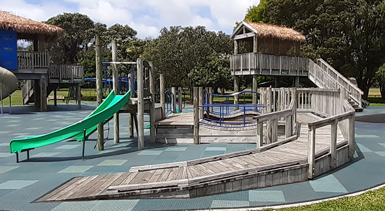 Ōrākei Domain - Ramp leading up into one of the wooden play huts with a green slide and the other huts and trees in the background.