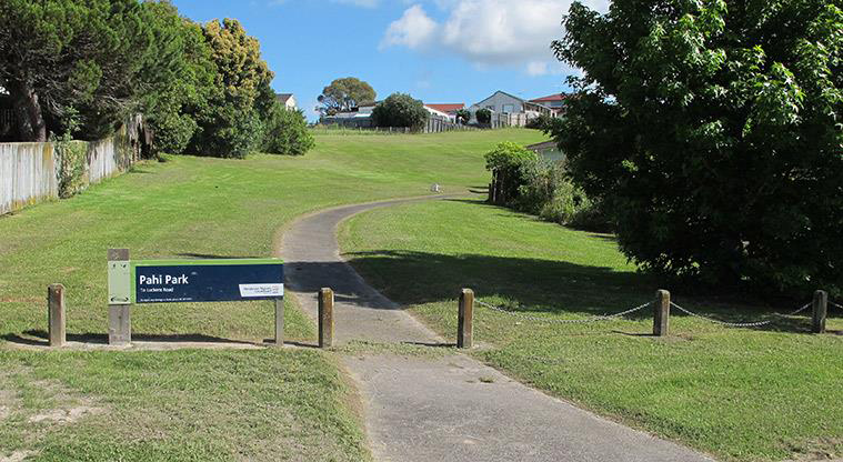 Pahi Park - West Harbour Drive entrance with open grassed space and a path running through the middle, and trees on both sides.