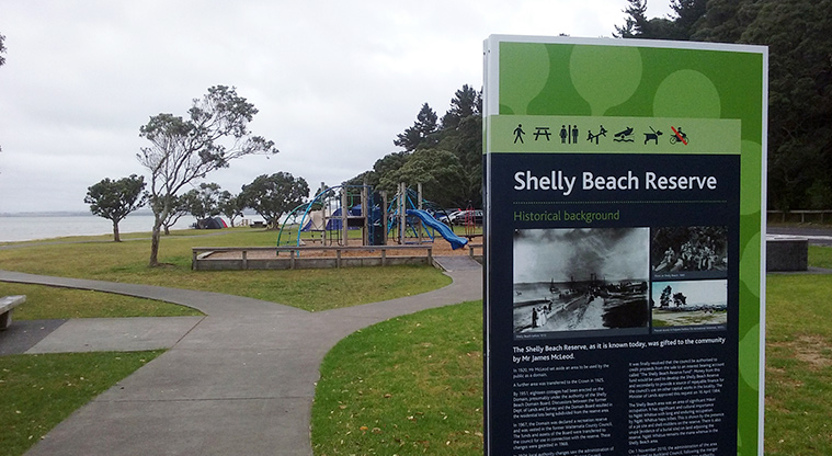 Shelly Beach Reserve - Information board showing the name and history of the reserve, with the playground and Kaipara Harbour in the background.