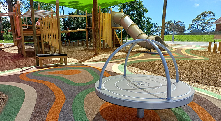 Shepherds Park - Grey spinning carousel on a colourful safety surface with the play structure in the background.