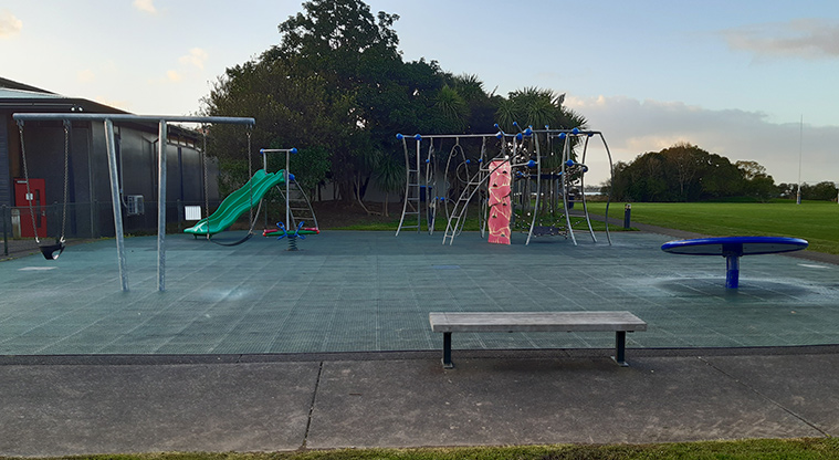 Shore Road Reserve - Wooden bench seat with the playground, sports fields and trees in the background.