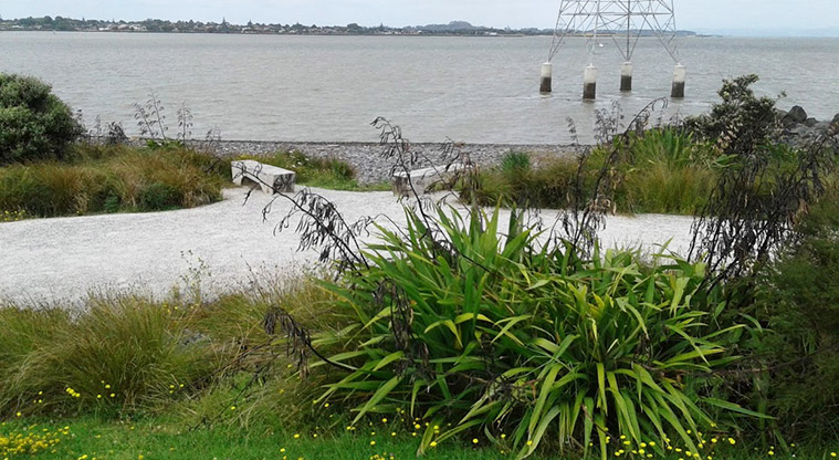 Taumanu Reserve - View from the reserve looking over a seating area and the foreshore, and across the Manukau Harbour to Mangere Bridge and Ambury Farm in the distance.