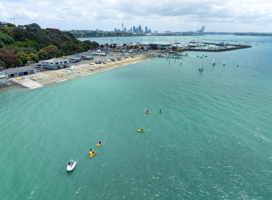 Aerial view of a bay with turquoise water, small boats and kayaks, marina buildings along the shore and Auckland’s skyline in the distance.
