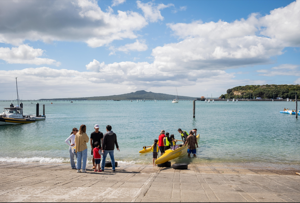 A group of people wade into the water with yellow kayaks on a boat ramp, with calm blue water, moored boats and a volcanic island covered with vegetation in the distance.