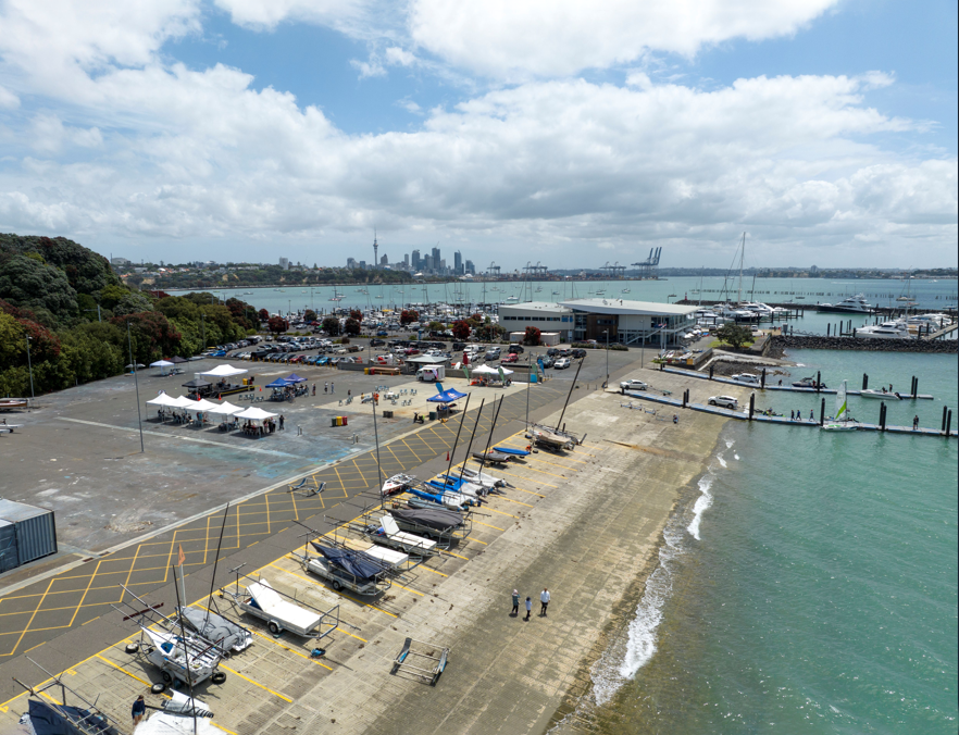 Small sailboats lined up on a ramp, with turquoise water, several docks extending into the harbour and Auckland’s skyline in the distance.