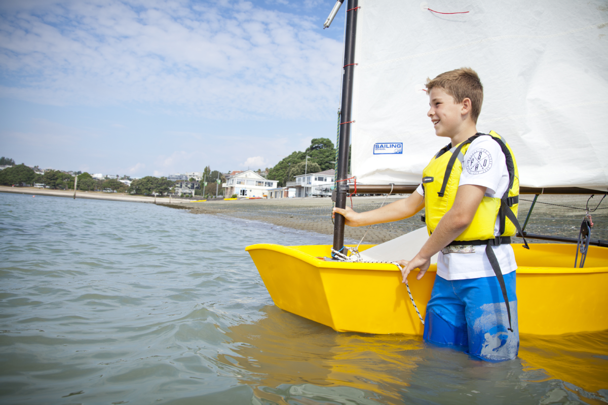 A young person wearing a lifejacket wades in the water holding onto a yellow dinghy.