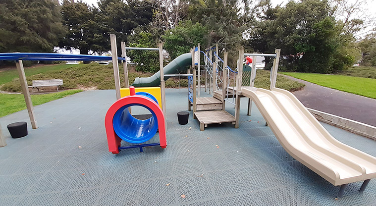 Waiatarua Reserve - Play structure with a double slide, colourful crawl tunnel, and platforms, surrounded by open grassed space and large trees.