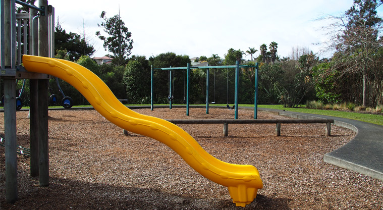 Xena Park - Close up of a yellow slide with swings in the background. Photo credit: Tracey Hodder.