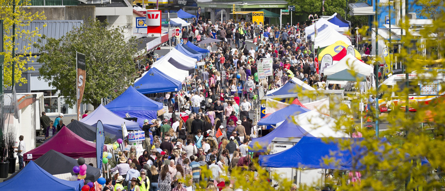 Photograph of Warkworth town centre during a community event, the streets are lined with marque market stalls and the road is full of people walking between stalls