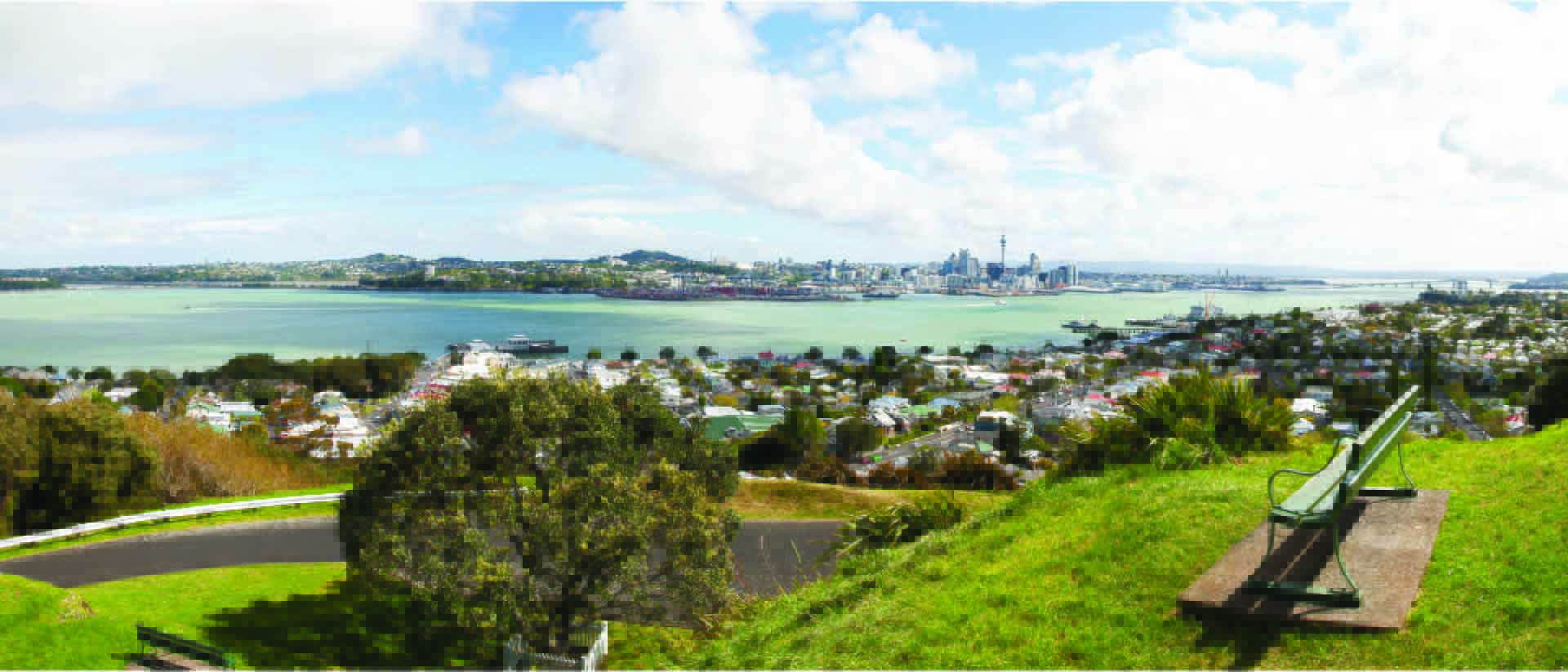 A bench on top of a grassy hill overlooking the Manukau Harbour with Auckland CBD in the background.