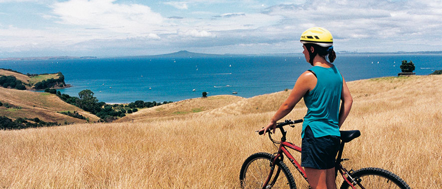 Women stands with bike while observing Rangitoto island from Shakespear Regional Park.