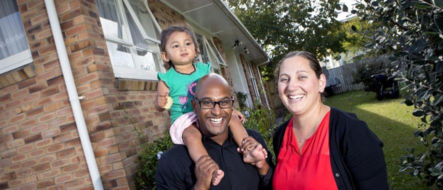 A smiling family of three in front of a brick home.