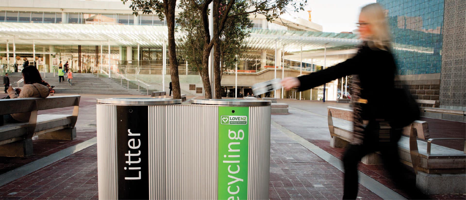 A person disposing of a plastic bottle into a recycling bin at an urban plaza.